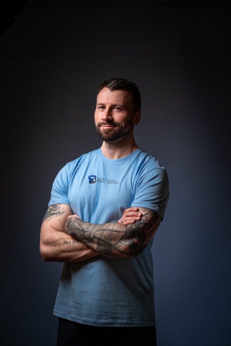 Patrick von Zeit für Fitness Mann mit muskulösen Armen, verschränkten Händen, in einem blauen Shirt vor neutralem Hintergrund.